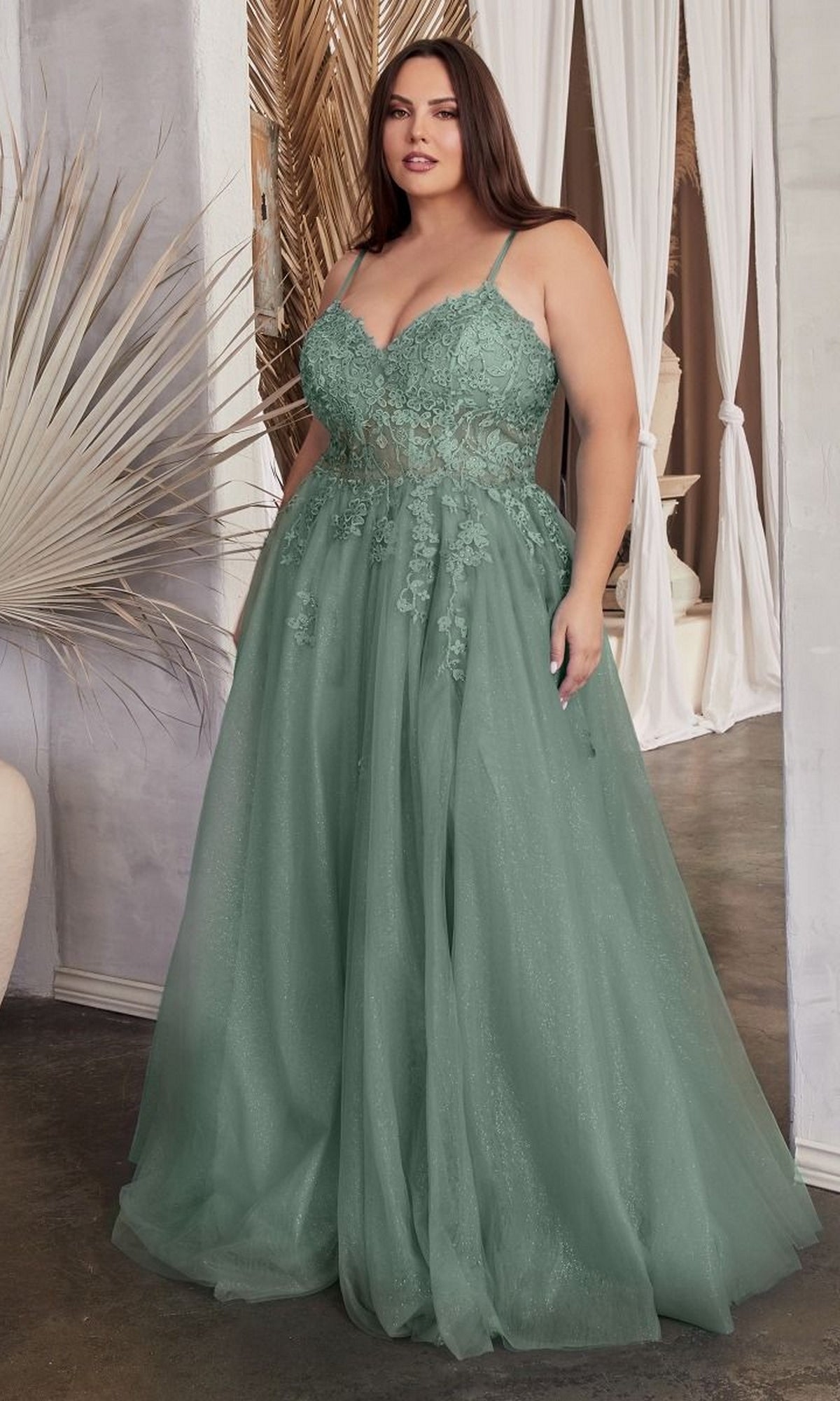 A woman in the Lace-Up Back Plus-Size Long Prom Ball Gown C148C, a floor-length sage green embroidered lace dress, stands indoors near neutral decor and draped curtains.